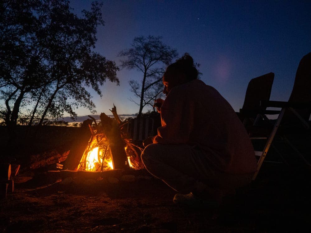 A person crouched by a campfire in a forest at night