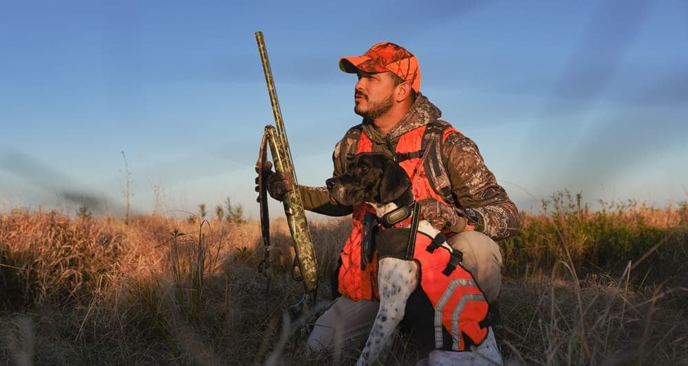 A hunter in camouflage with a dog in a meadow at sunrise