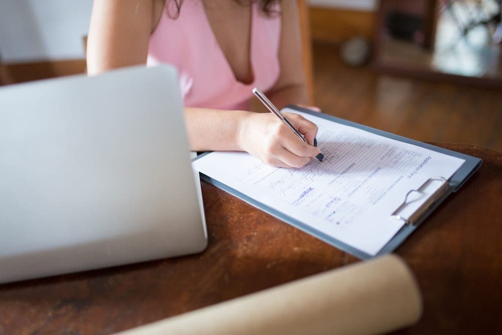 A person filling out a form on a clipboard next to a laptop