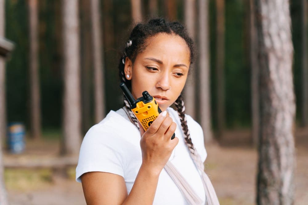 A woman talking into a yellow handheld radio in a pine forest