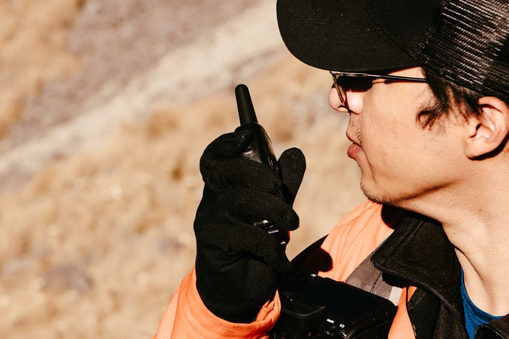 A man in gloves talking into a handheld radio on a dry hillside